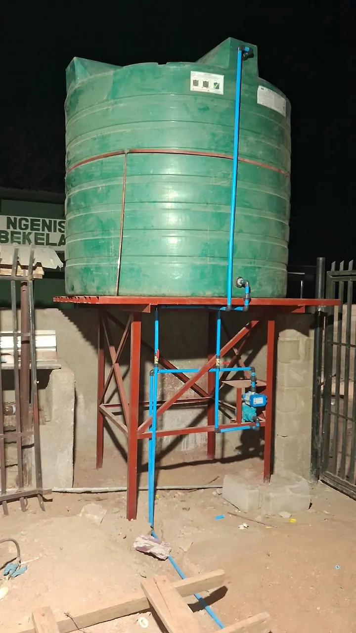 Green water storage tank on a steel stand with blue supply pipework, valves and a pump installed underneath